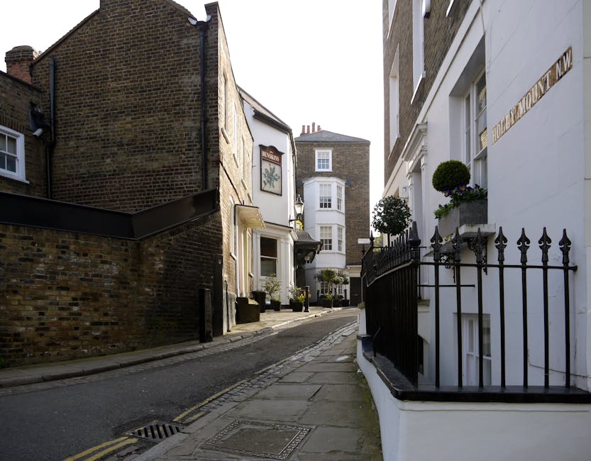 A narrow residential street with cobblestone paving, lined with closely spaced terraced houses featuring white and red brick facades. Several potted plants and small trees in containers are positioned