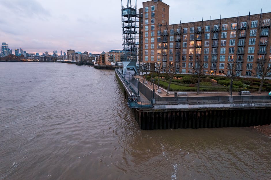 Aerial view of a riverside residential area during dusk, showing a large brick building with multiple floors and numerous windows near the water's edge. To the right, there is a paved promenade with b