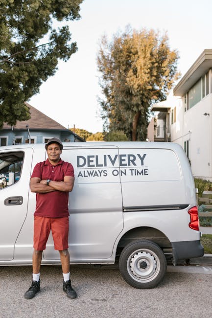 A man wearing a white t-shirt, jeans, white sneakers, and a black cap is loading cardboard boxes into the side door of a white delivery van parked on the street in front of residential houses with pit